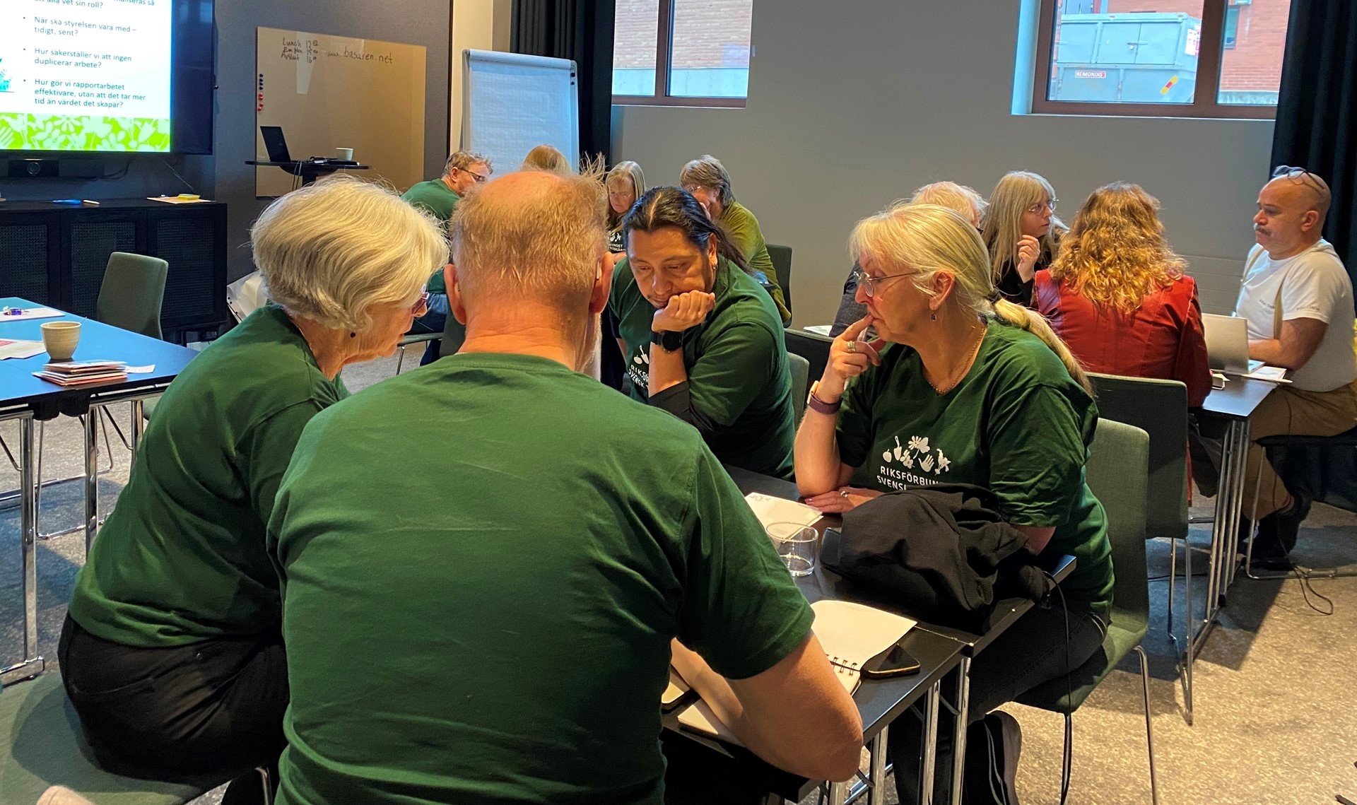 Grupparbete i ett konferensrum 12 personer i olika åldrar placerade på tre olika bord, är försjunkna i diskutioner. Till vänster i bild är en bildskärm, tavla och antexkningsblock.